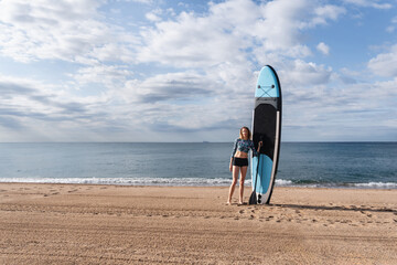Young woman holding stand up paddle board on a sandy beach at seaside, enjoying summer vacation...