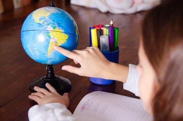 A young girl pointing at a globe and doing her homework on a wooden table, surrounded by school supplies. 