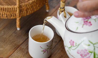 Hot tea being poured from a floral teapot into a matching cup on a wooden table. Warm beverage serving, Asian tea culture, relaxation, hospitality, traditional drink, and healthy lifestyle concept