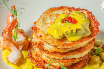 Crispy potato pancakes stacked with creamy yellow curry sauce, garnished with shrimp, herbs, and...
