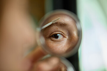 Close-up of woman eye applying brow gel seen through mirror with focused expression. Concept of...