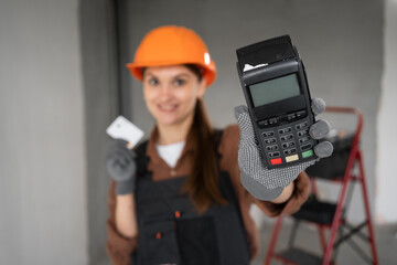 Woman builder in uniform holding POS terminal and credit card, looking at camera. Concept of...