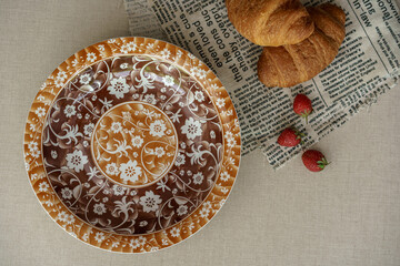 Vintage style breakfast flat lay. An empty ornate floral plate in brown tones next to fresh croissants and strawberries on a newspaper.