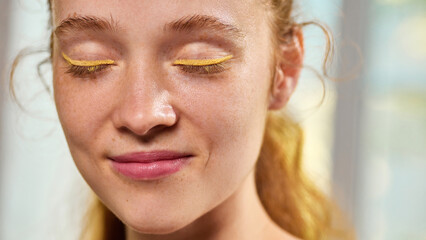 Freckled woman smiling with eyes closed wearing bright eyeliner on eyelids. Concept of creativity,...
