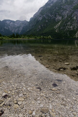 A crystal-clear lake reveals its rocky bed, reflecting the majestic, tree-covered mountains under a soft sky.