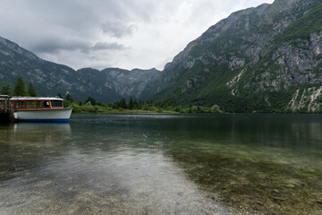 A classic wooden boat rests peacefully on a clear lake, surrounded by towering, lush green mountains under a dramatic sky.