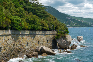 A historic stone wall meets the vibrant blue Adriatic Sea, with lush green trees crowning the hillside above, under a cloudy sky.