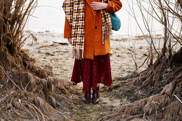 A woman in orange coat stands between dry branches by a sandy shore.