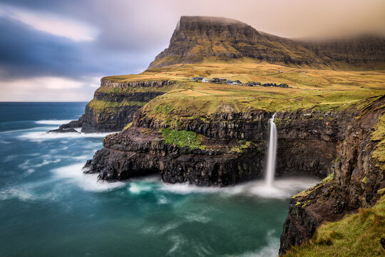 View of cascading waterfall plunging into the turquoise sea below dramatic cliffs topped with green grass and small village, Gásadalur, Faroe Islands. - Powered by Adobe