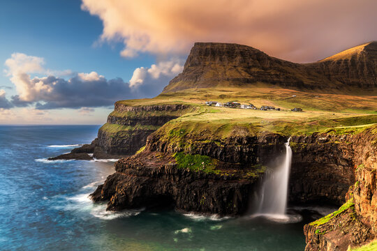 View of cascading waterfall plunges from a cliffside village into the turbulent sea, under a sky painted with dawn's first light, Gasadalur, Faroe Islands.