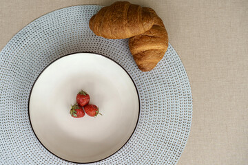 Minimalist breakfast flat lay. Top view of three fresh strawberries on a white plate with croissants on a blue placemat. Healthy eating concept.