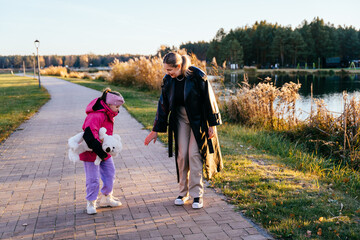 Girl with Down syndrome walking with mother near lake holding teddy bear