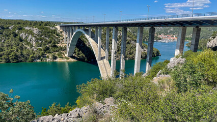 Scenic arch bridge spanning turquoise river with lush green hills and marina in the background under a clear blue sky, showcasing travel infrastructure and natural beauty