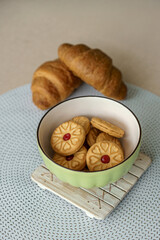 Teatime still life featuring a green bowl of jam-filled shortbread cookies and freshly baked croissants on a beige background.