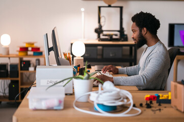 An African American man works diligently at his computer in a modern office. He is focused on the screen, typing on the keyboard. The workspace is organized with various tech items.