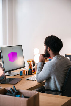 An African American man sits at his desk, intently viewing a computer screen showing a 3D purple cube. He is focused on a design or modeling project.