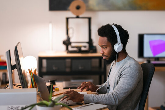An African American man wearing headphones is engrossed in work on his laptop at a contemporary desk. He is focused on his digital tasks in a well-equipped home office.