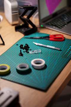 An African American person's workspace with a cutting mat, craft knife, scissors, and tape rolls. They are engaged in a detailed project, with a laptop nearby.