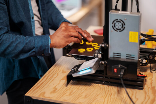 An African American person is carefully removing yellow 3D printed objects from the print bed of a 3D printer. This highlights the hands-on process of additive manufacturing.
