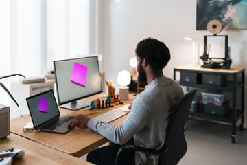 An African American man works on 3D design at his home office. He uses a laptop and monitor displaying a 3D model, with a 3D printer nearby. He is focused on his creative work.