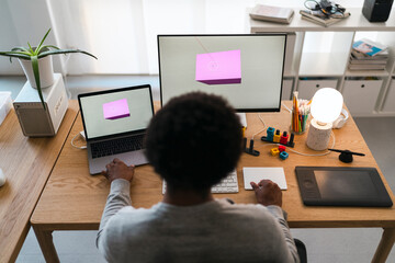 An African American designer works on a 3D model using a dual-screen setup with a laptop and monitor. They are focused on their creative project at a well-lit home office desk.