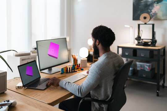 An African American man works on 3D design at his home office. He uses a laptop and monitor displaying a 3D model, with a 3D printer nearby. He is focused on his creative work.