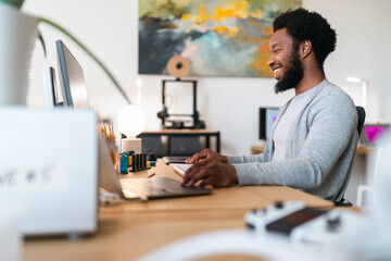 An African American man smiles broadly while working on his laptop and computer monitor at a bright, modern office desk. He is focused and engaged in his professional tasks.
