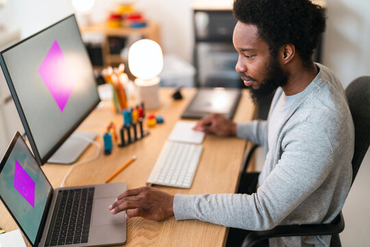 An African American man works on graphic design at his home office desk. He uses a laptop and an external monitor, both displaying a vibrant pink geometric shape.