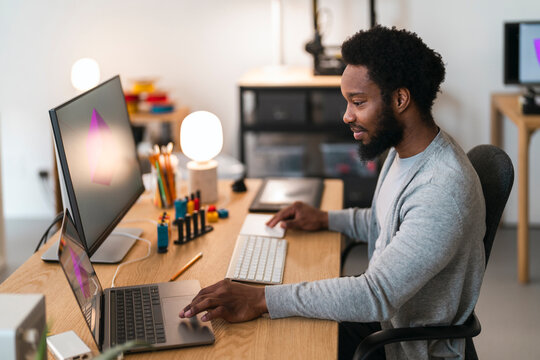 An African American man works at his desk, focused on his computer setup. He uses a laptop and an external monitor displaying a 3D graphic, engaged in creative work.