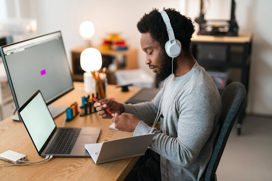 An African American man with headphones sits at a desk, engaged in work. He holds a red pen, with a laptop and large monitor visible, indicating a productive environment.