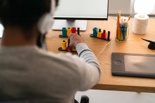 An African American person with headphones works at a desk, adjusting a creative control panel. A computer monitor and graphics tablet are visible, suggesting a digital design or editing task.