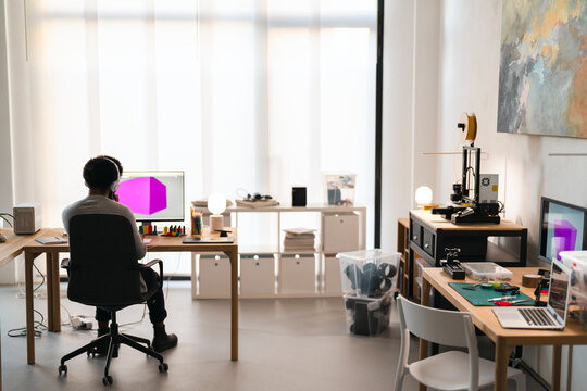An African American person sits at a desk, wearing headphones and working on a computer displaying a 3D model. A 3D printer is set up in their bright, modern office.