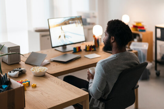 An African American man sits at a desk, holding a cup and looking at a computer monitor. A bowl of popcorn and a game controller are on the wooden desk.