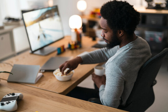 An African American man sits at a desk, eating popcorn and holding a cup. He is focused on the computer monitor, with a game controller nearby, enjoying his leisure time.