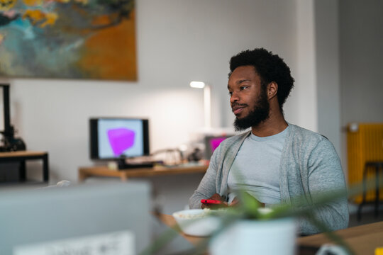An African American man sits at a desk, looking away, holding his phone. A laptop and a bowl are on the desk. A computer screen with a pink cube is visible in the background.