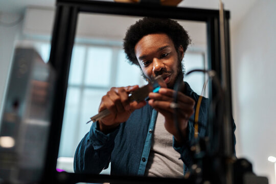 An African American man uses a caliper to precisely measure a component. He is focused on his work with a 3D printer, demonstrating engineering skills.