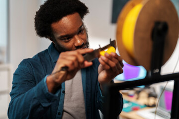 An African American man precisely measures yellow 3D printer filament with a caliper. He focuses intently on the task, ensuring accuracy for his engineering or design project.