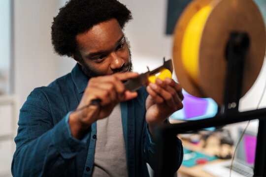 An African American man precisely measures yellow 3D printer filament with a caliper. He focuses intently on the task, ensuring accuracy for his engineering or design project.
