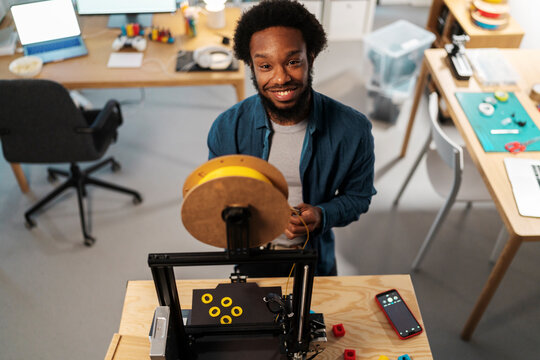 An African American man smiles at the camera while setting up a 3D printer. He is feeding yellow filament into the machine, with yellow printed objects on the bed.