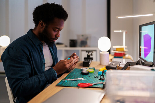 An African American man meticulously works on a small blue object at his desk. Surrounded by tools and components, he engages in a focused creative or engineering project.