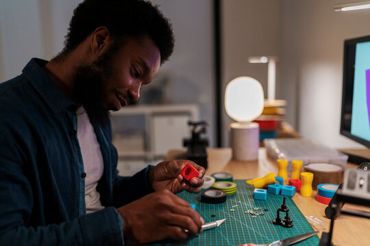 An African American man is focused on a crafting project at his desk. He holds a red 3D-printed part and a knife, surrounded by tools and components. He is engaged in a creative hobby.