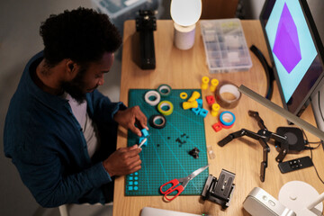 An African American man is focused on a crafting project at his desk. He uses a tool to assemble small blue components on a green mat, surrounded by various supplies and a computer monitor.