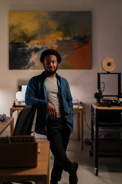 An African American man with an afro and beard stands confidently in his home office, leaning on a chair. He smiles at the camera, surrounded by creative tools like a 3D printer.