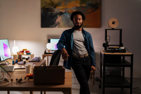 An African American man stands confidently in his creative workspace, leaning on a chair. He looks at the camera, surrounded by design tools, a computer with a 3D model, and a 3D printer.