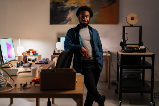 An African American man stands confidently in his creative workspace, leaning on a chair. His desk is filled with design tools, a laptop, and a 3D printer is visible in the background