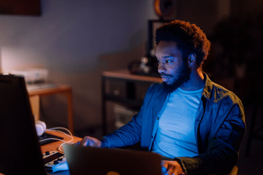 An African American man is focused on his laptop screen in a dimly lit room. Blue light illuminates his face as he works or plays. A game controller is nearby.
