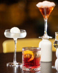 A richly colored red cocktail with a vibrant orange peel garnish is presented in a textured rocks glass, surrounded by other sophisticated drinks in a dimly lit bar setting.