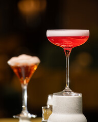 A richly colored red cocktail with a vibrant orange peel garnish is presented in a textured rocks glass, surrounded by other sophisticated drinks in a dimly lit bar setting.