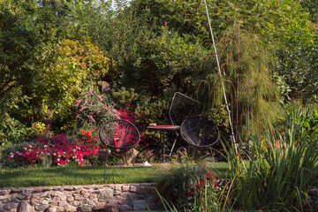 A fisherman's place in a decorative garden by a pond. Two chairs and a table in the middle, next to a fishing rod and a net for catching fish; blooming flowers, decorative shrubs.