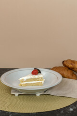 Slice of layered vanilla cake with white frosting, garnished with a strawberry and cookie. Sweet dessert on a plate with croissants in the background.
